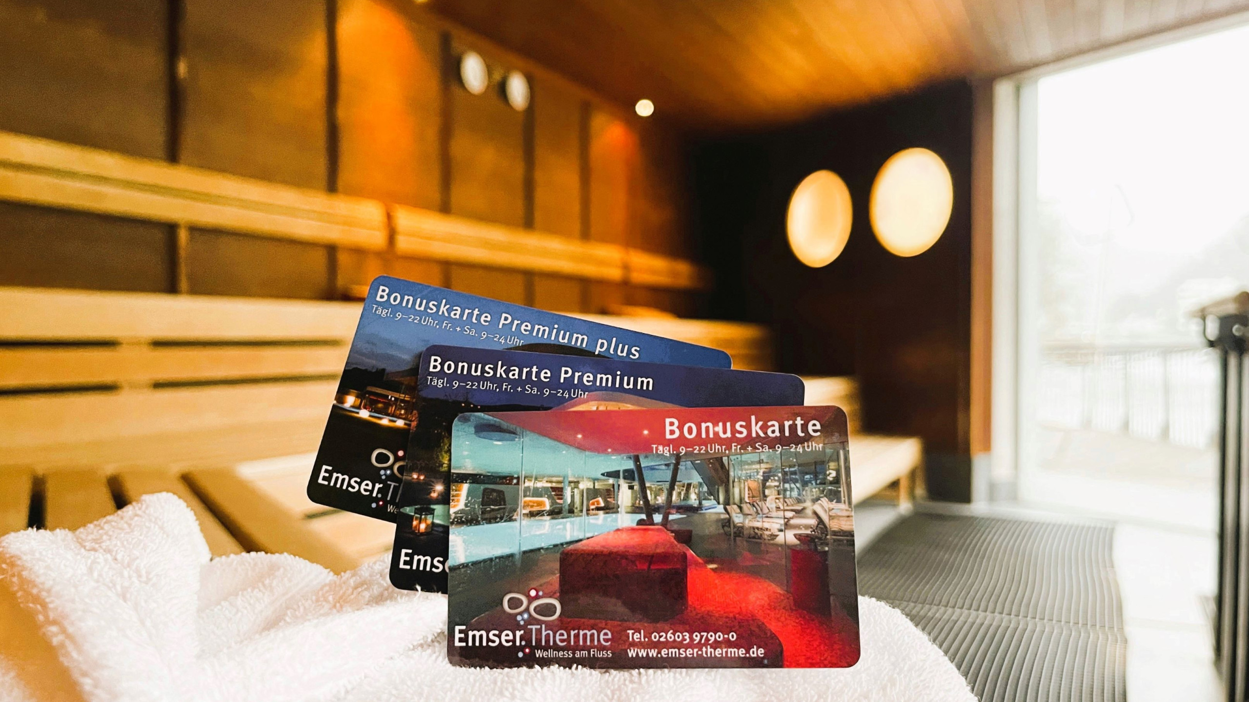 Drei Bonuskarten der Emser Therme liegen auf einem weißen Handtuch, vor einem stilvollen Saunainterior mit Holzverkleidung und einem großen Fenster mit Ausblick.