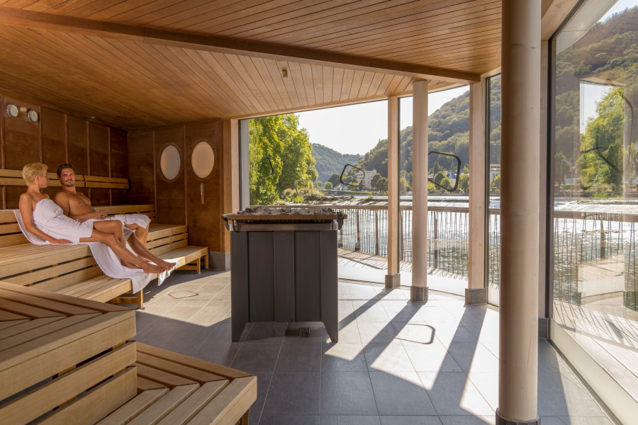 Ein Paar entspannt in einer modernen Sauna mit Holzbankverkleidung. Durch große Fenster blickt es auf eine malerische Landschaft mit Wasser und Bäumen. Sanfte Lichtakzente schaffen eine gemütliche Atm