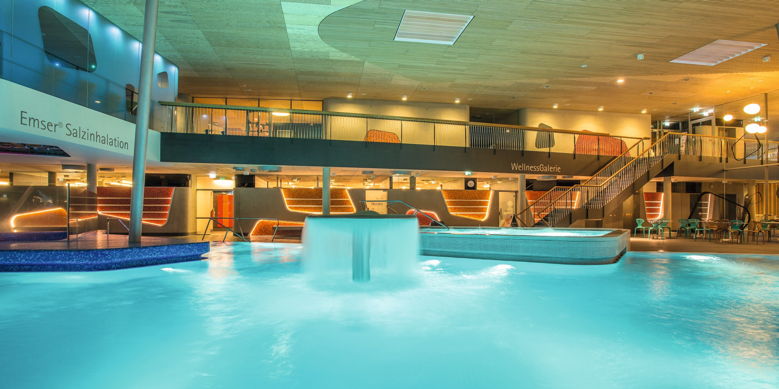 Innenansicht eines modernen Wellnessbereichs mit einem beheizten Pool, einer Wasserfontäne und stilvollem Mobiliar. Warmes Licht und Holzdetails schaffen eine entspannende Atmosphäre.