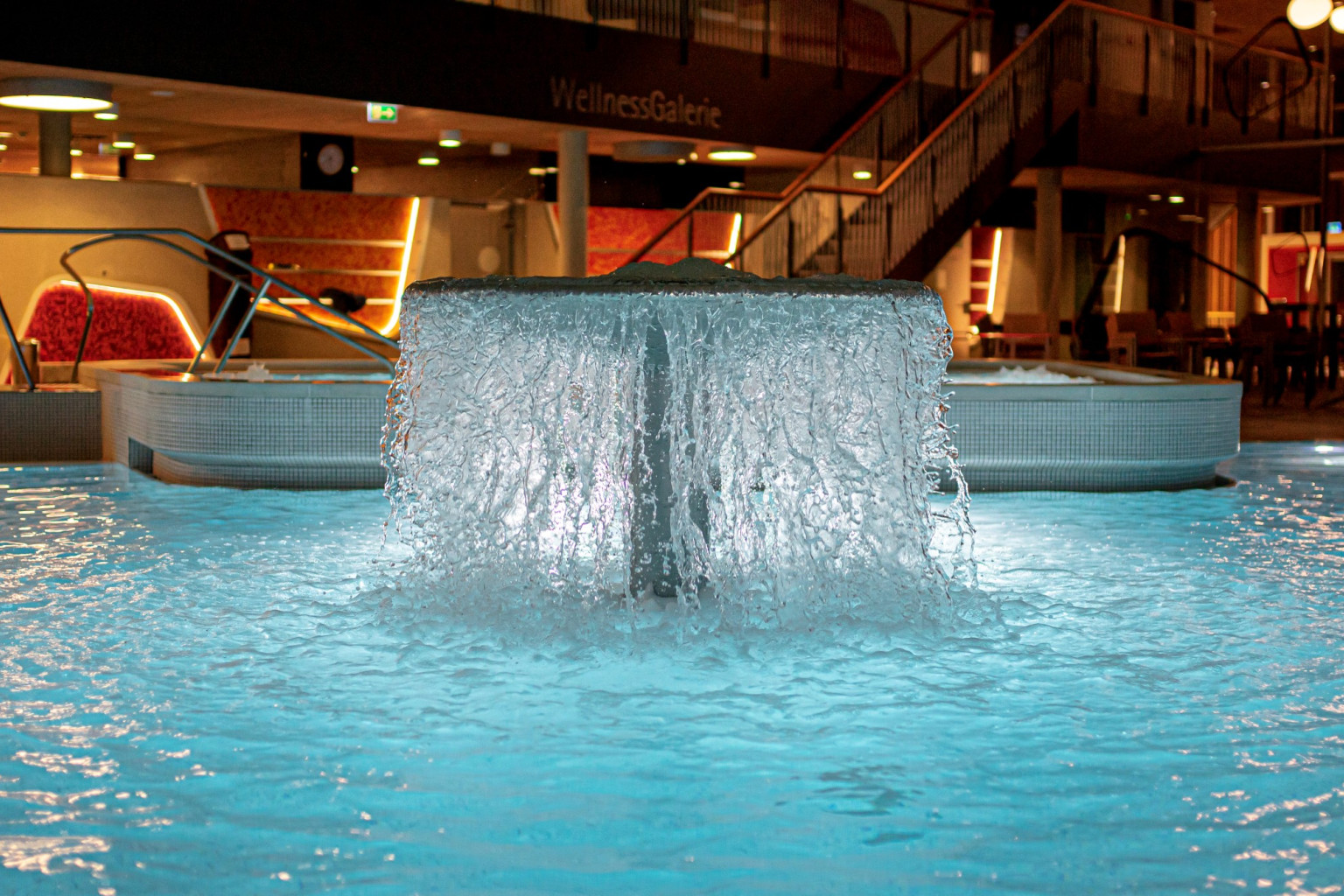 Ein modernes Wellnessbereich mit einem sprudelnden Wasserfall in einem klaren, blauen Pool. Die stilvolle Beleuchtung schafft eine entspannende Atmosphäre, während im Hintergrund ein gemütlicher Sitzb