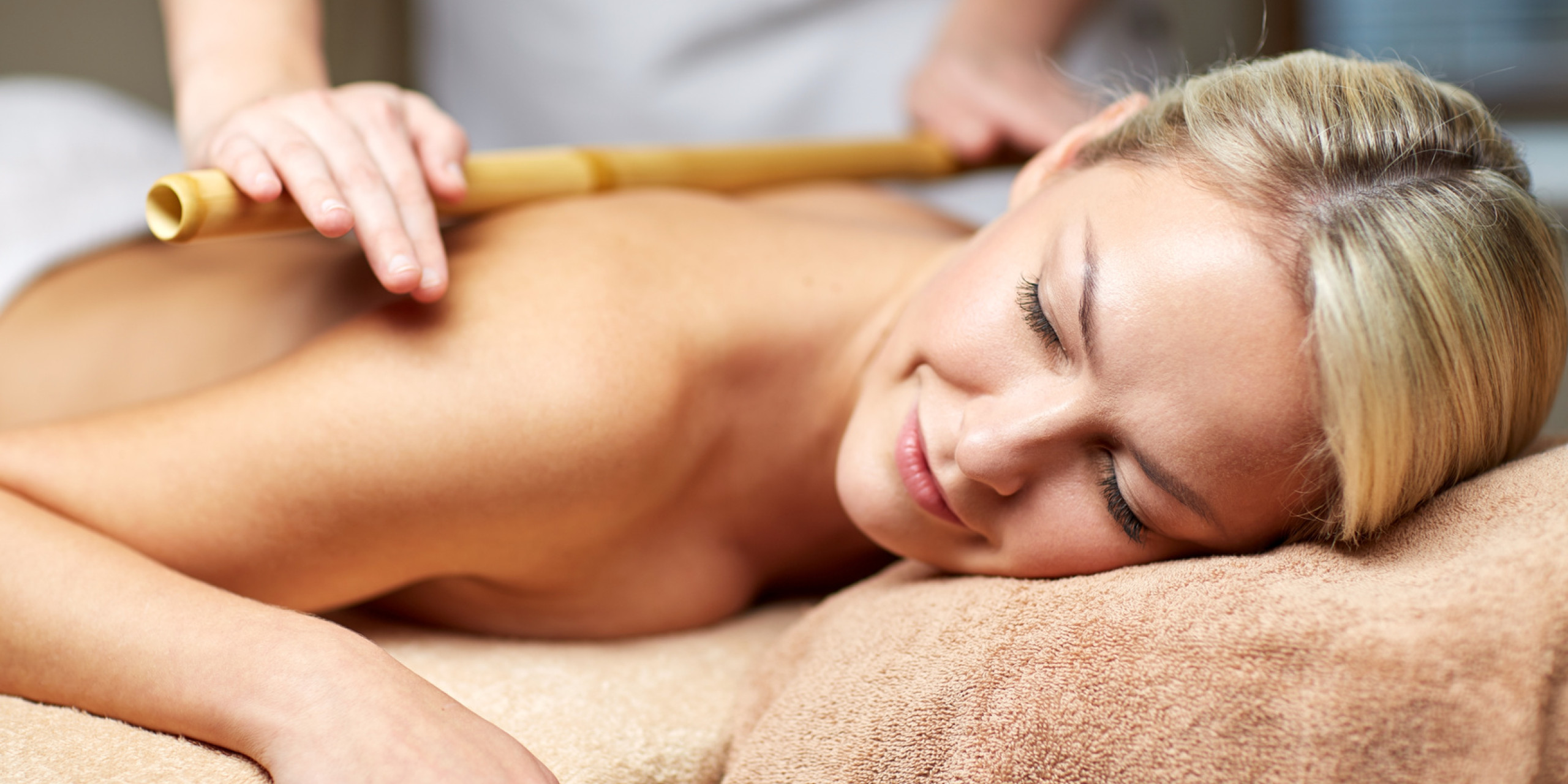 Eine Frau liegt entspannt auf einer Liege, während eine Therapeutin eine Massage mit einem Bambusstab durchführt. Die Atmosphäre wirkt beruhigend und wohltuend, mit sanften, warmen Farben.