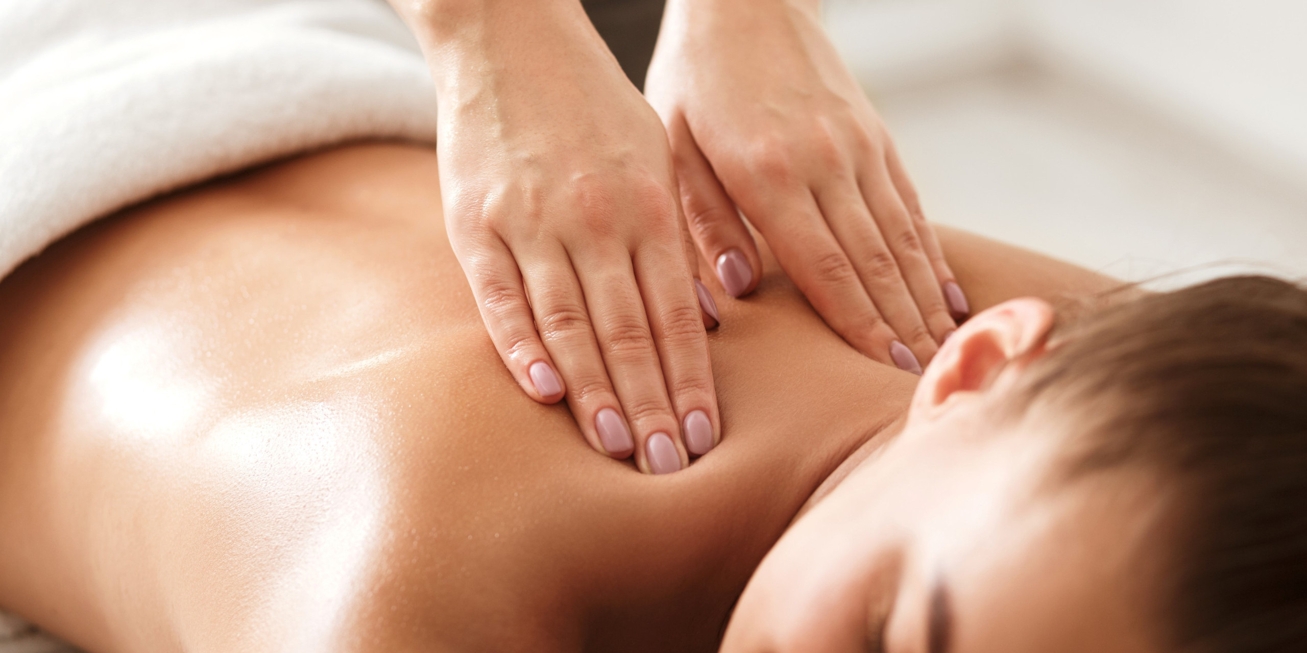 Eine Person erhält eine entspannende Rückenmassage. Die Hände der Masseurin sind sanft auf der Haut platziert, die von einem weißen Handtuch umgeben ist. Die Atmosphäre wirkt beruhigend und friedlich.