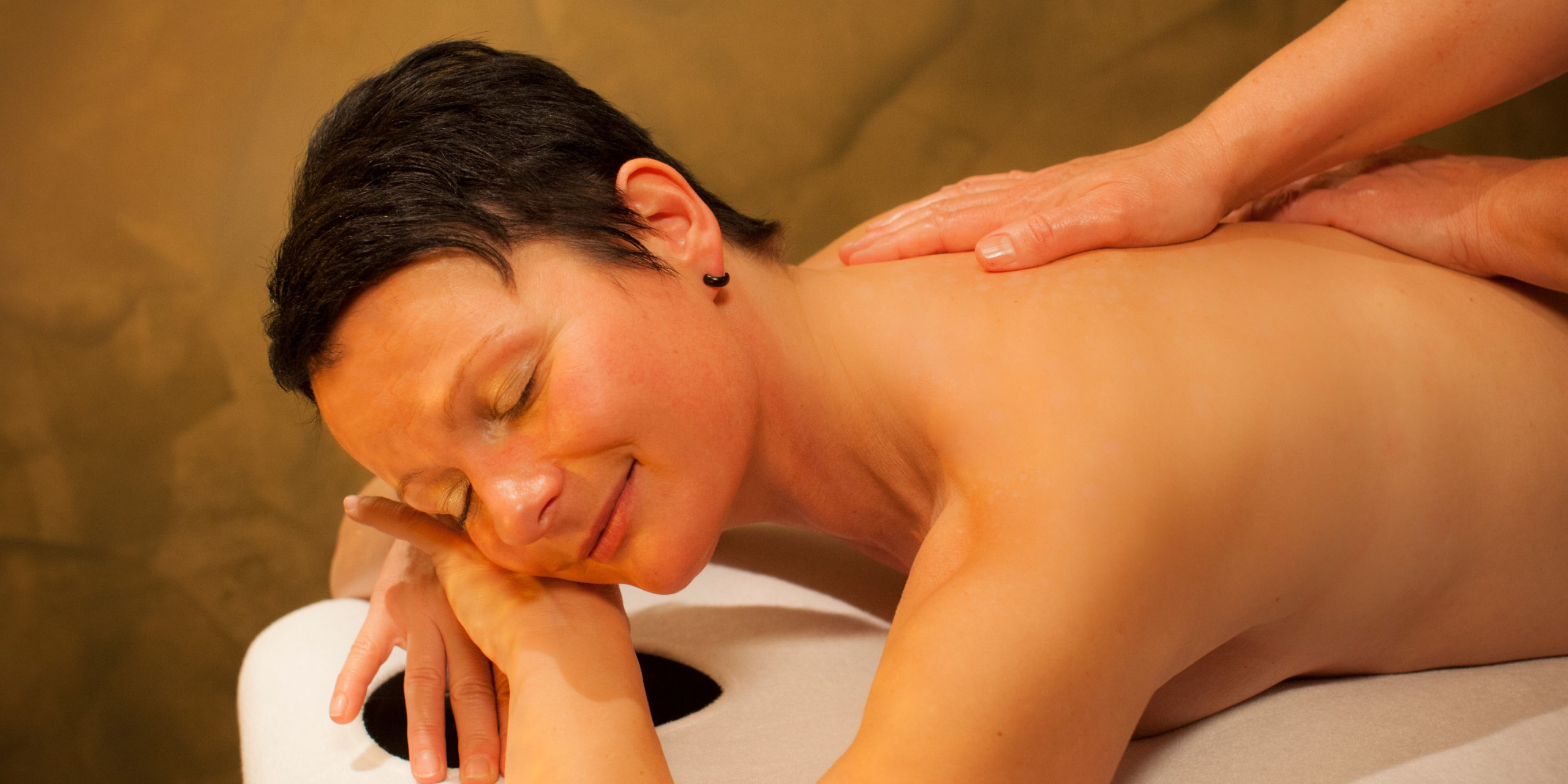 Eine entspannte Frau liegt auf einer Massageliege. Sie hat geschlossene Augen und lächelt sanft, während eine Hand ihren Rücken massiert. Der Hintergrund ist in warmen Brauntönen gehalten.