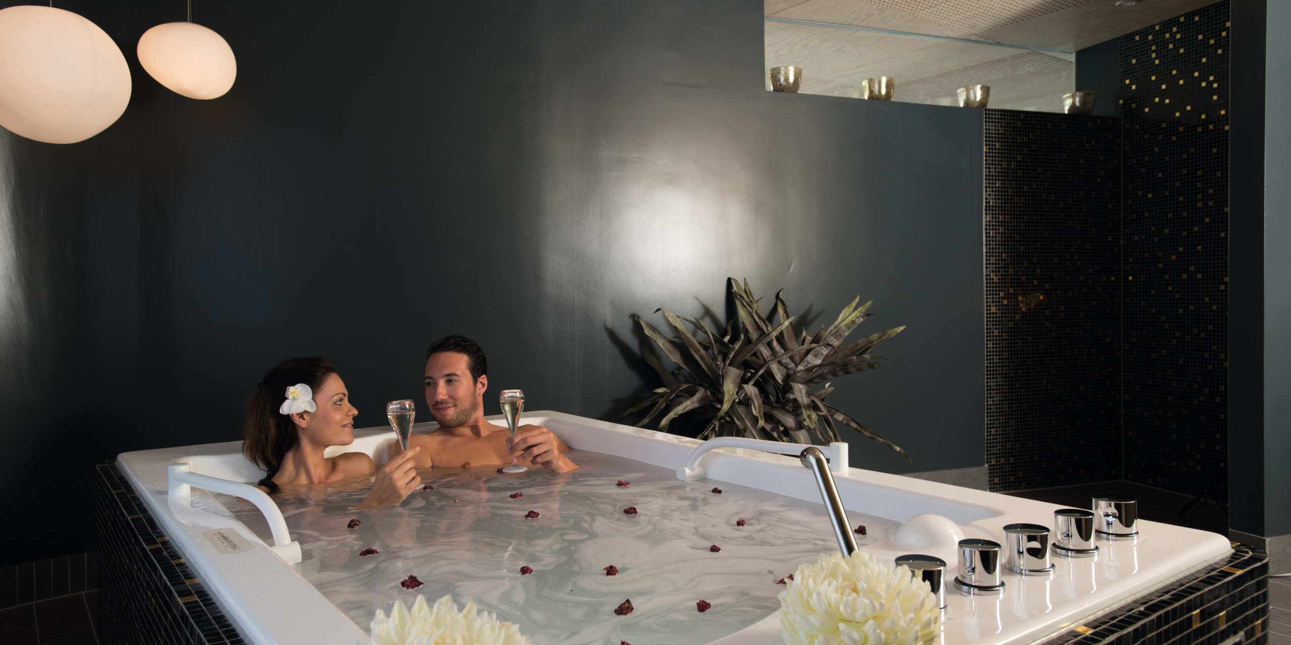 Ein Paar entspannt in einem modernen Whirlpool, umgeben von dunklen Wänden. Sie halten gläserne Drinks und lächeln sich an, während Rosenblätter auf dem Wasser schwimmen. Pflanzen dekorieren den Raum.