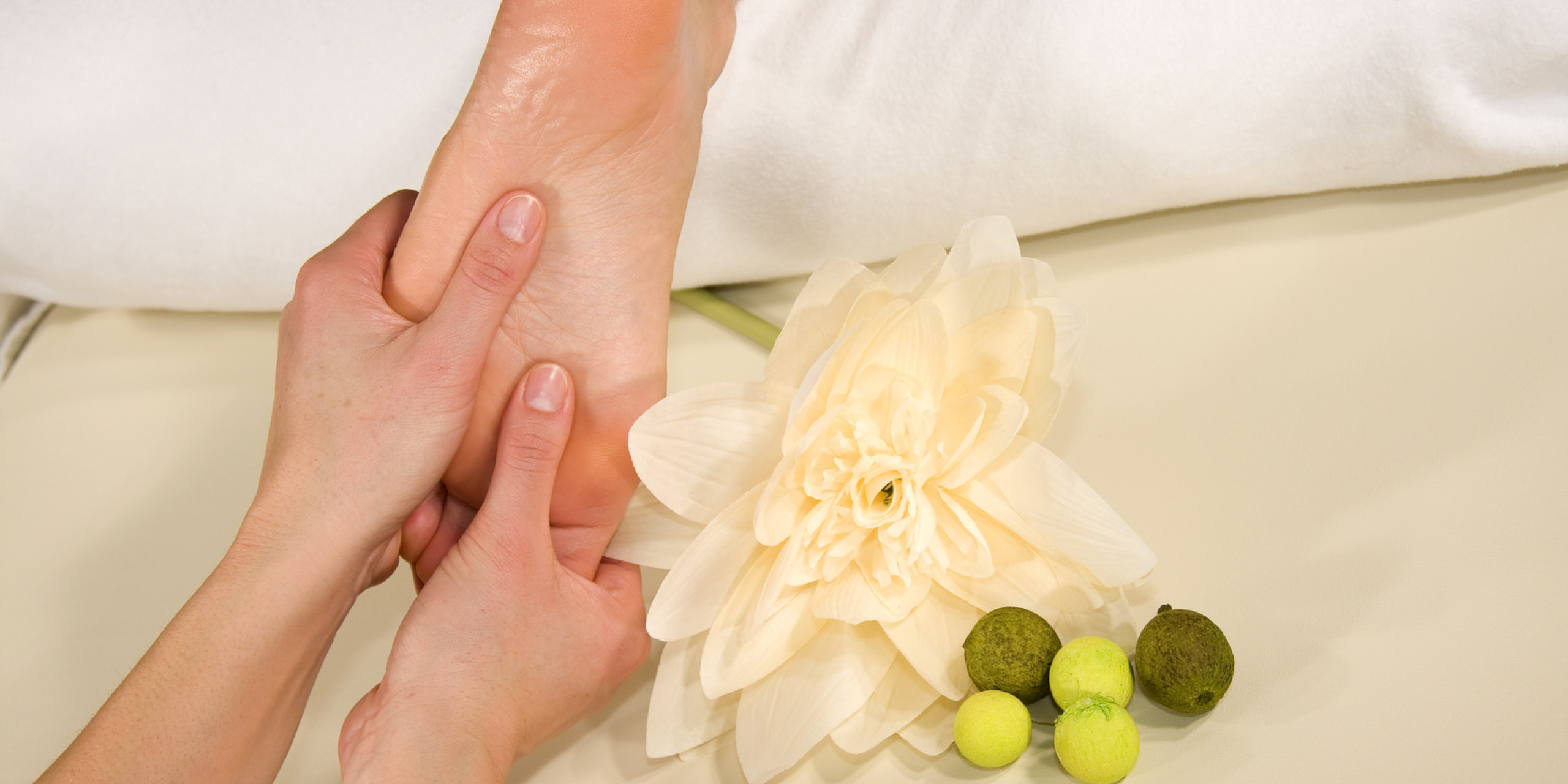 Eine Hand massiert sanft einen Fuß, umgeben von einem weißen Kissen und einer großen, cremefarbenen Blume. Neben dem Fuß liegen grüne und braune kleine Objekte. Entspannung und Pflege stehen im Vorder