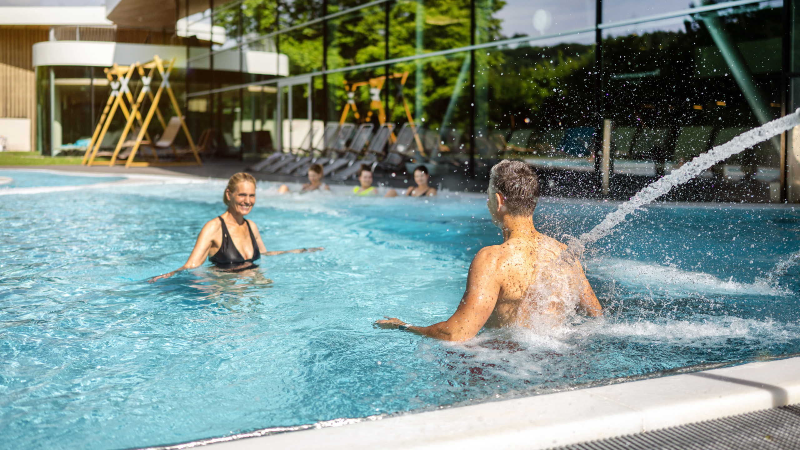 Zwei Personen genießen einen sonnigen Tag im Pool. Eine Frau in einem schwarzen Bikini lacht, während ein Mann im Wasser spritzt. Im Hintergrund sind Liegen und eine moderne Architektur zu sehen.
