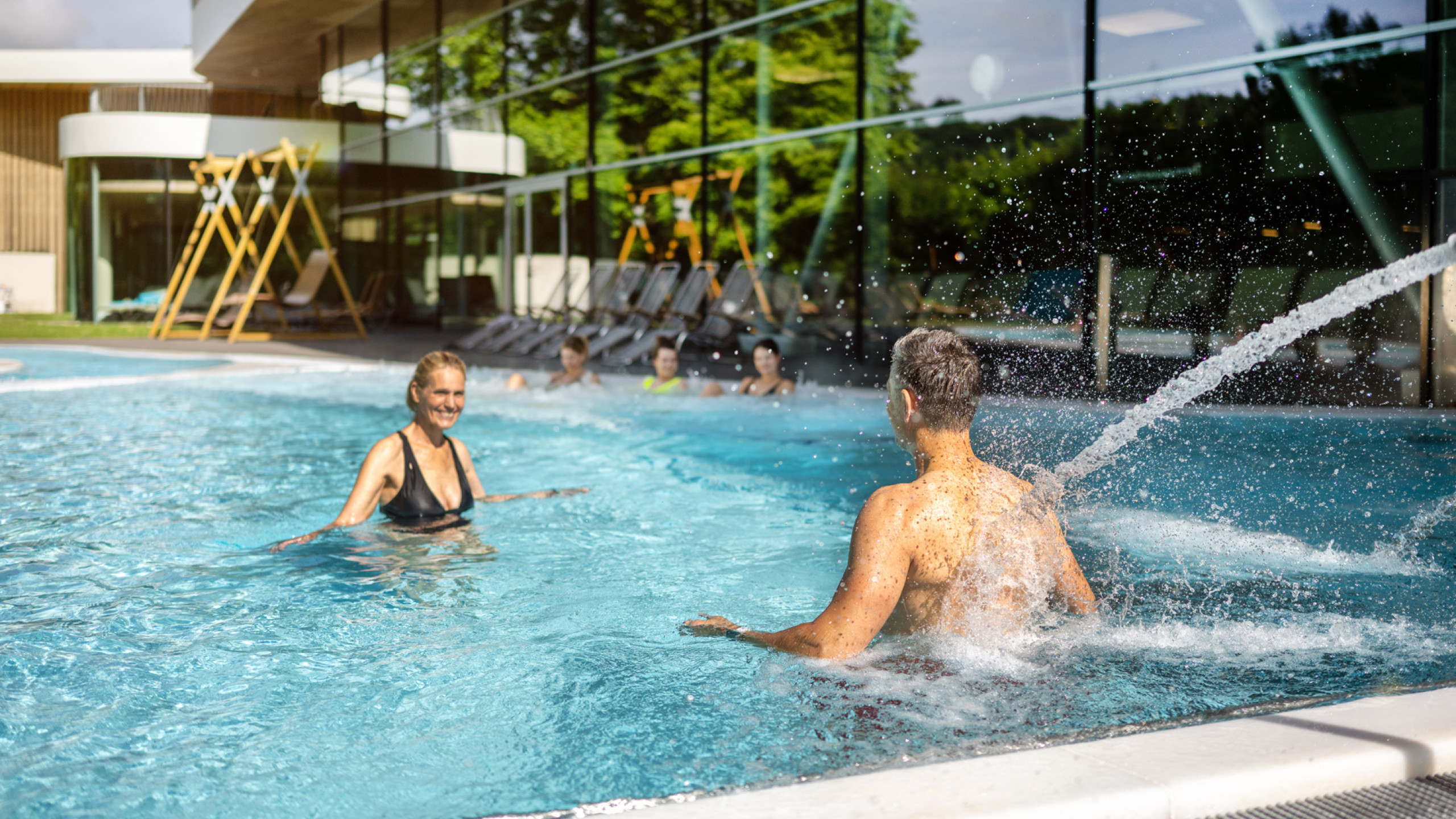 Zwei Personen genießen einen sonnigen Tag im Pool. Eine Frau in einem schwarzen Bikini lacht, während ein Mann im Wasser spritzt. Im Hintergrund sind Liegen und eine moderne Architektur zu sehen.